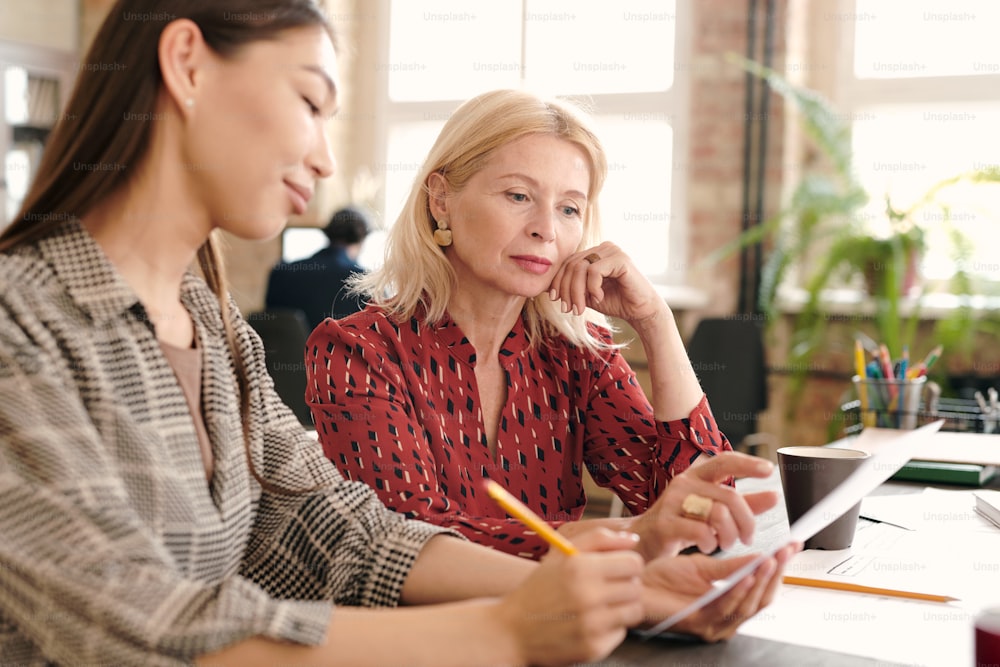 Young and mature serious female designers looking at sketch on paper while one of them pointing at it during discussion of details at meeting