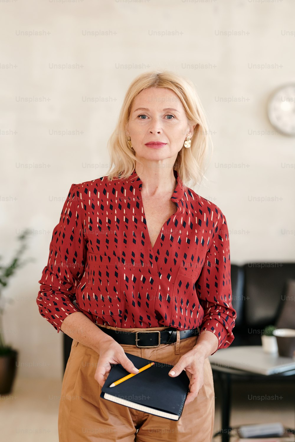 Mature confident businesswoman in smart red blouse and beige pants holding notebook and pencil while standing in office environment