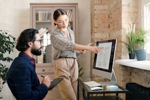 Young female mobile application developer with tablet pointing at sketch on computer screen while discussing it with her male colleague