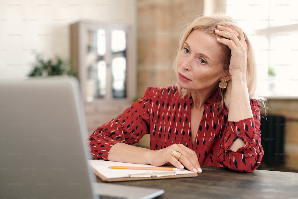 Concentrated mature blond businesswoman sitting in front of laptop and looking at online data on display during network in office environment