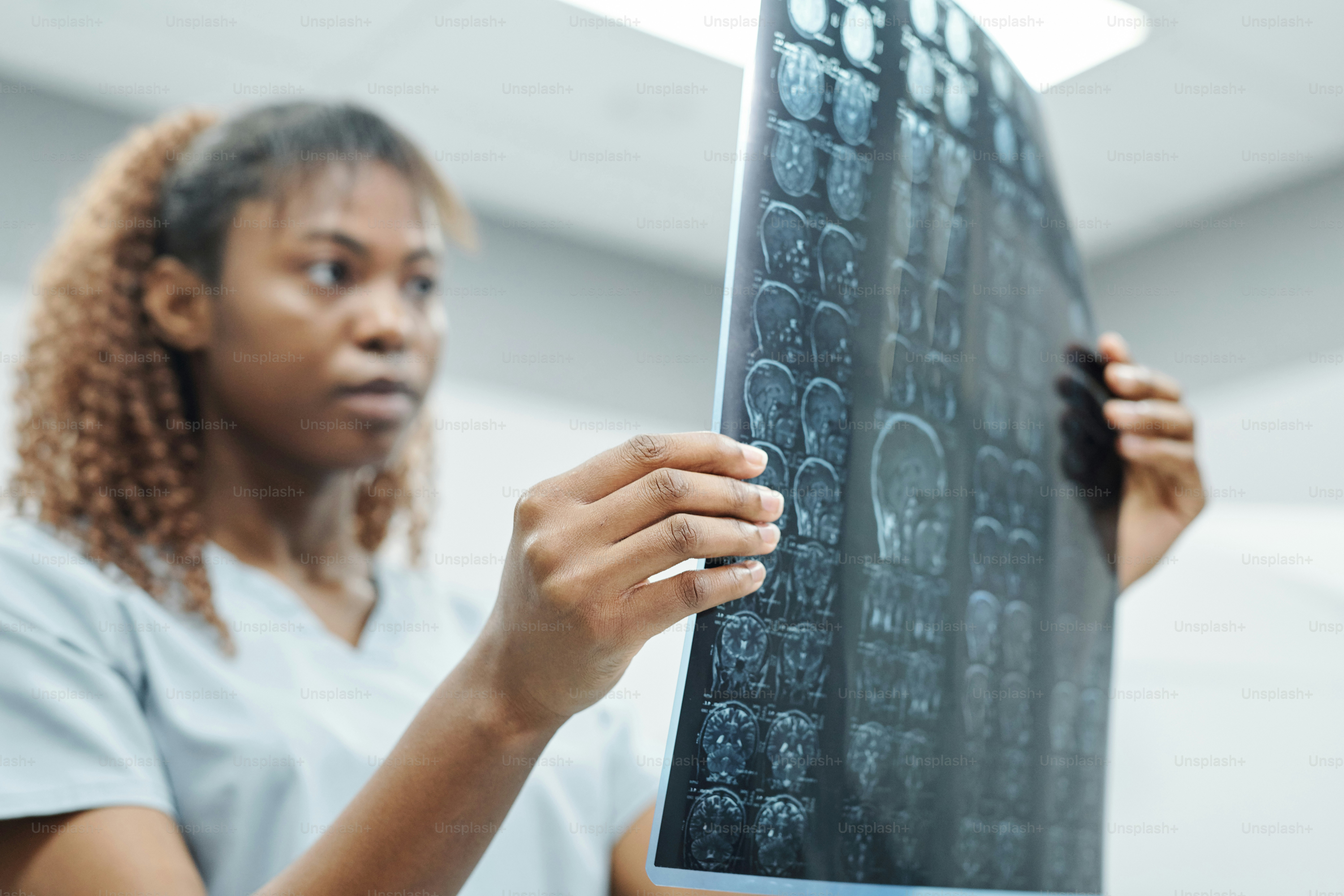Hand of young African female radiologist in uniform analyzing x-ray image of patient head against environment of modern medical office