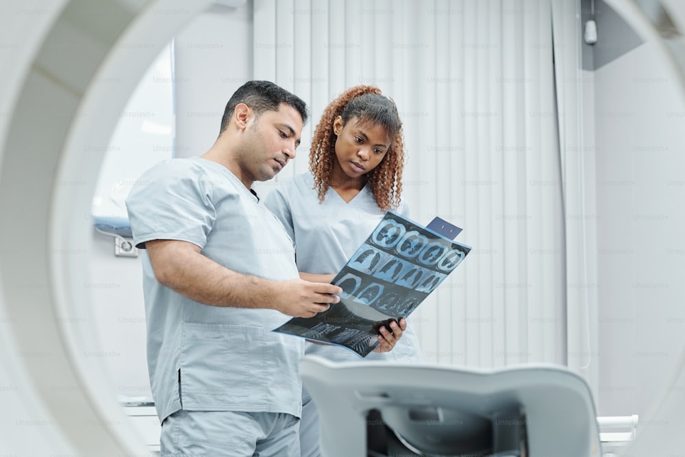 Serious male radiologist and his African assistant in uniform analyzing x-ray image of patient in modern medical office by ultra sound equipment