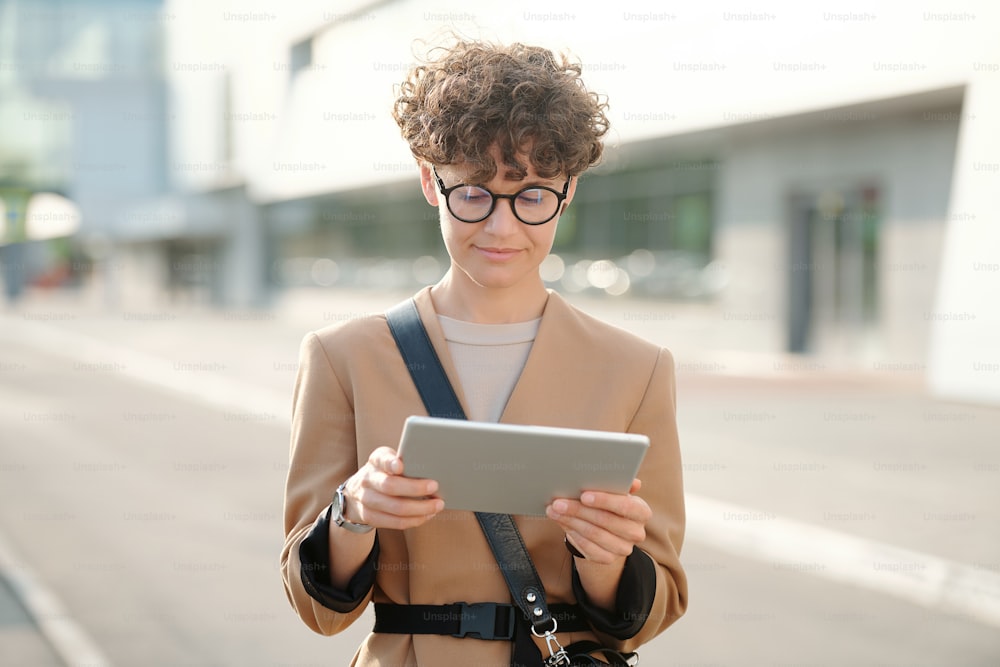 Junge hübsche brünette Geschäftsfrau mit digitalem Tablet, die sich ein Business-Webinar oder eine Konferenz gegen die Straße in einer städtischen Umgebung ansieht