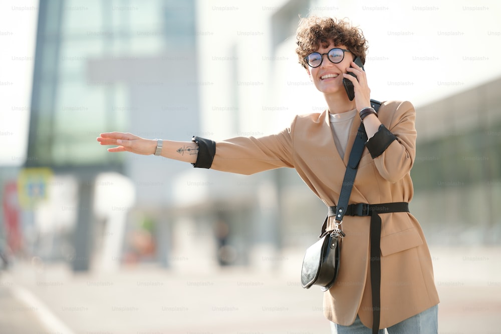 Jovem mulher elegante sorridente com cliente de consultoria de smartphone no telefone enquanto está de pé pela estrada e pegando táxi no centro da cidade