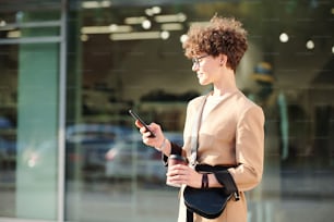 Young curly brunette businesswoman with drink and handbag messaging in smartphone while standing against large shop window of mall