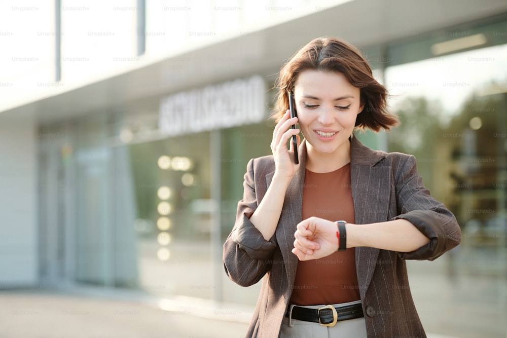 Jovem empresária elegante contemporânea com cliente de consultoria de smartphone no telefone ao ar livre e olhando para o wistwatch pela estrada