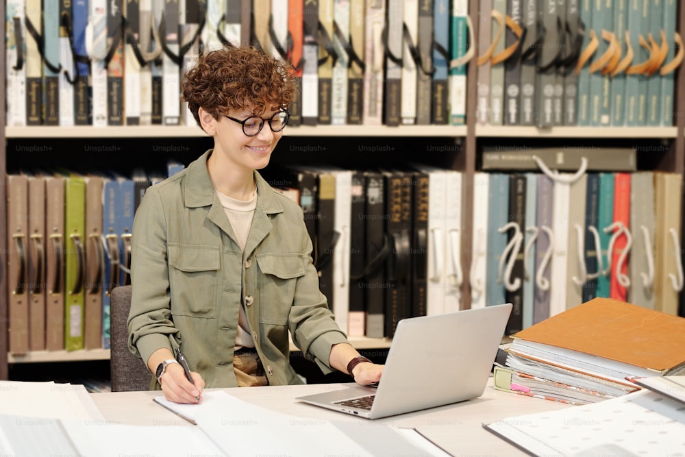 인테리어 디자인 스튜디오의 젊은 갈색 머리 여성 매니저가 노트북 디스플레이를 보면서 테이블별로 새로운 구색에 대해 메모하고 있다