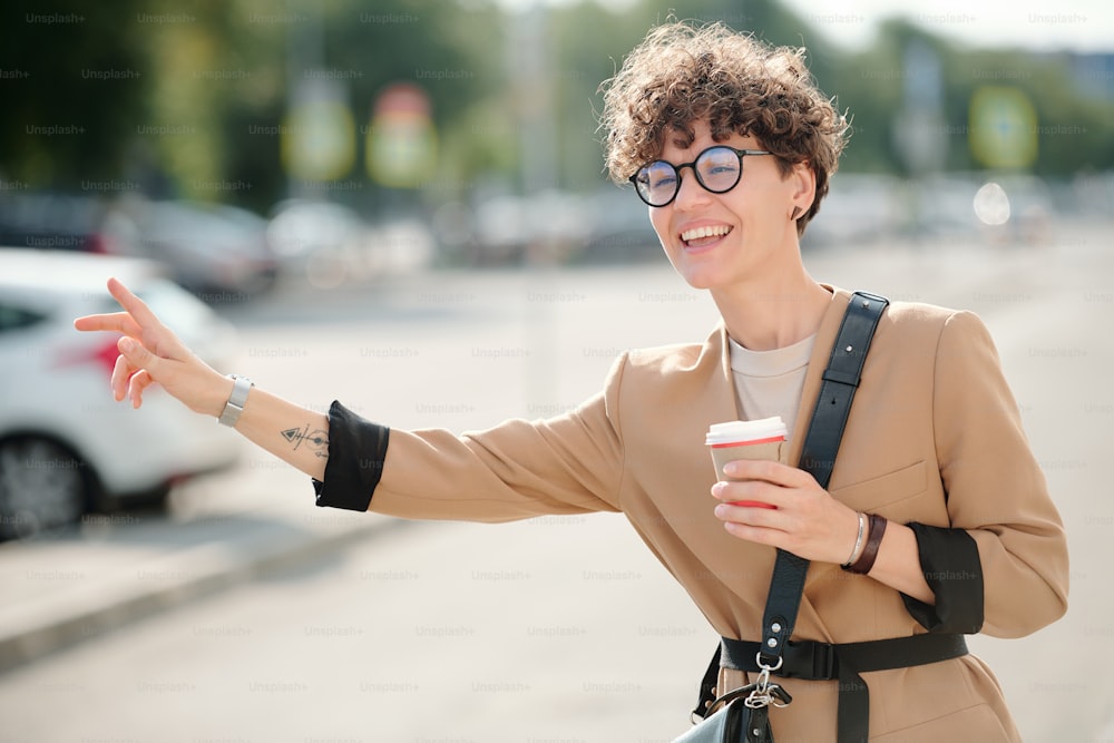 Young cheerful businesswoman with glass of coffee standing by road and catching taxi against modern building in urban environment