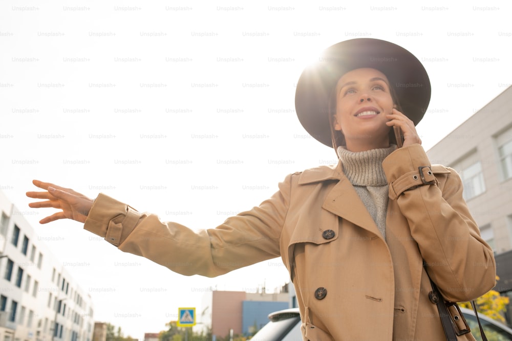 Giovane donna d'affari sorridente elegante in trench beige e cappello nero che cerca di prendere un taxi mentre si trova su strada in ambiente urbano