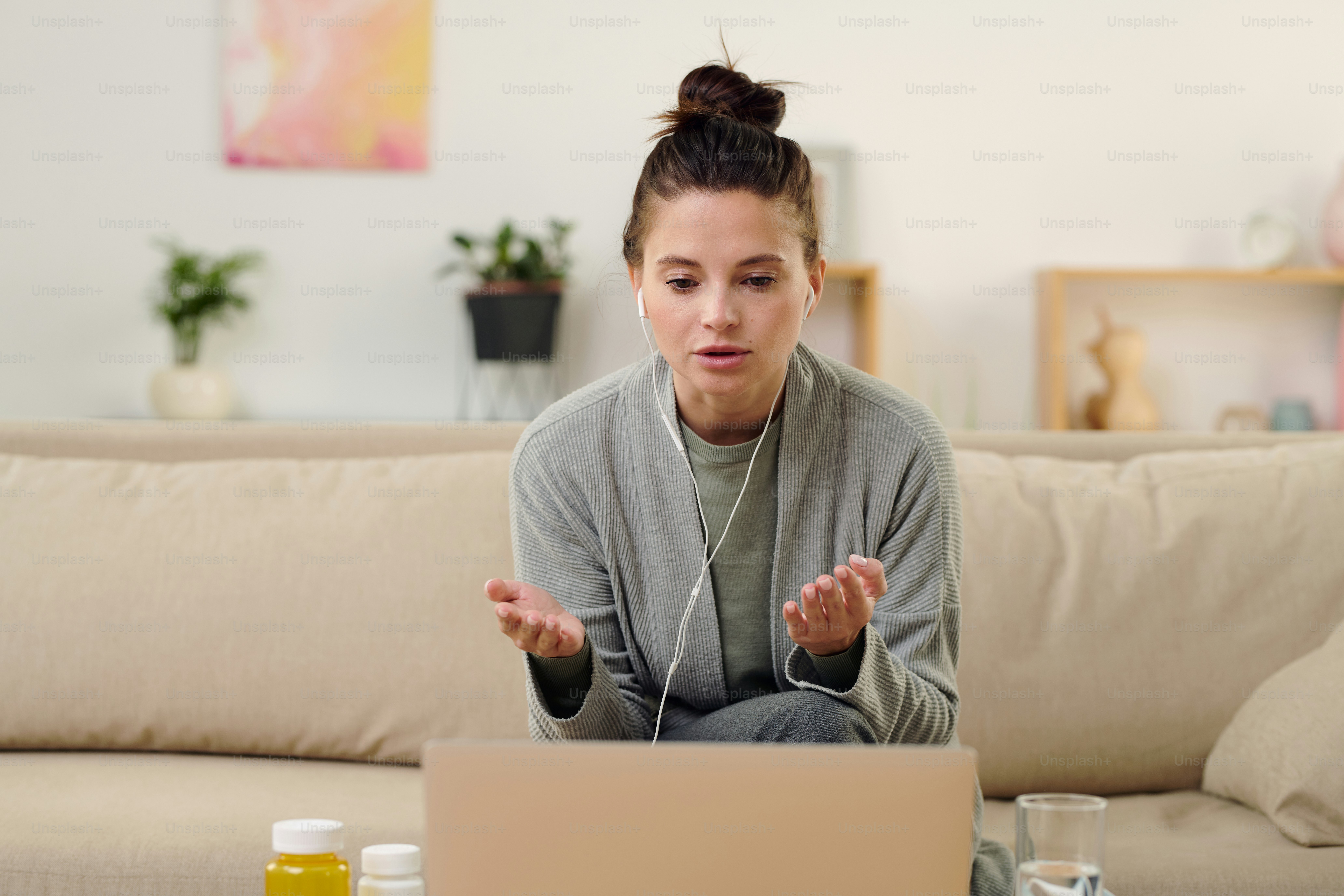 Junge brünette Frau mit Kopfhörern erklärt dem Online-Arzt ihr Problem, während sie auf der Couch vor dem Laptop in der häuslichen Umgebung sitzt