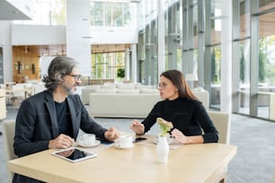 Dois colegas confiantes em roupas casuais inteligentes e óculos discutindo notícias financeiras ou projetos de sua empresa por xícara de café