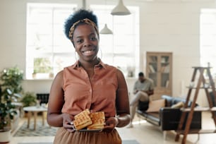 Jovem mulher africana feliz com waffles caseiros olhando para você com sorriso dentuço enquanto está de pé contra seu marido sentado no sofá