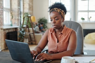 Serious young African female freelancer in casualwear working in front of laptop while sitting in armchair by table in home environment