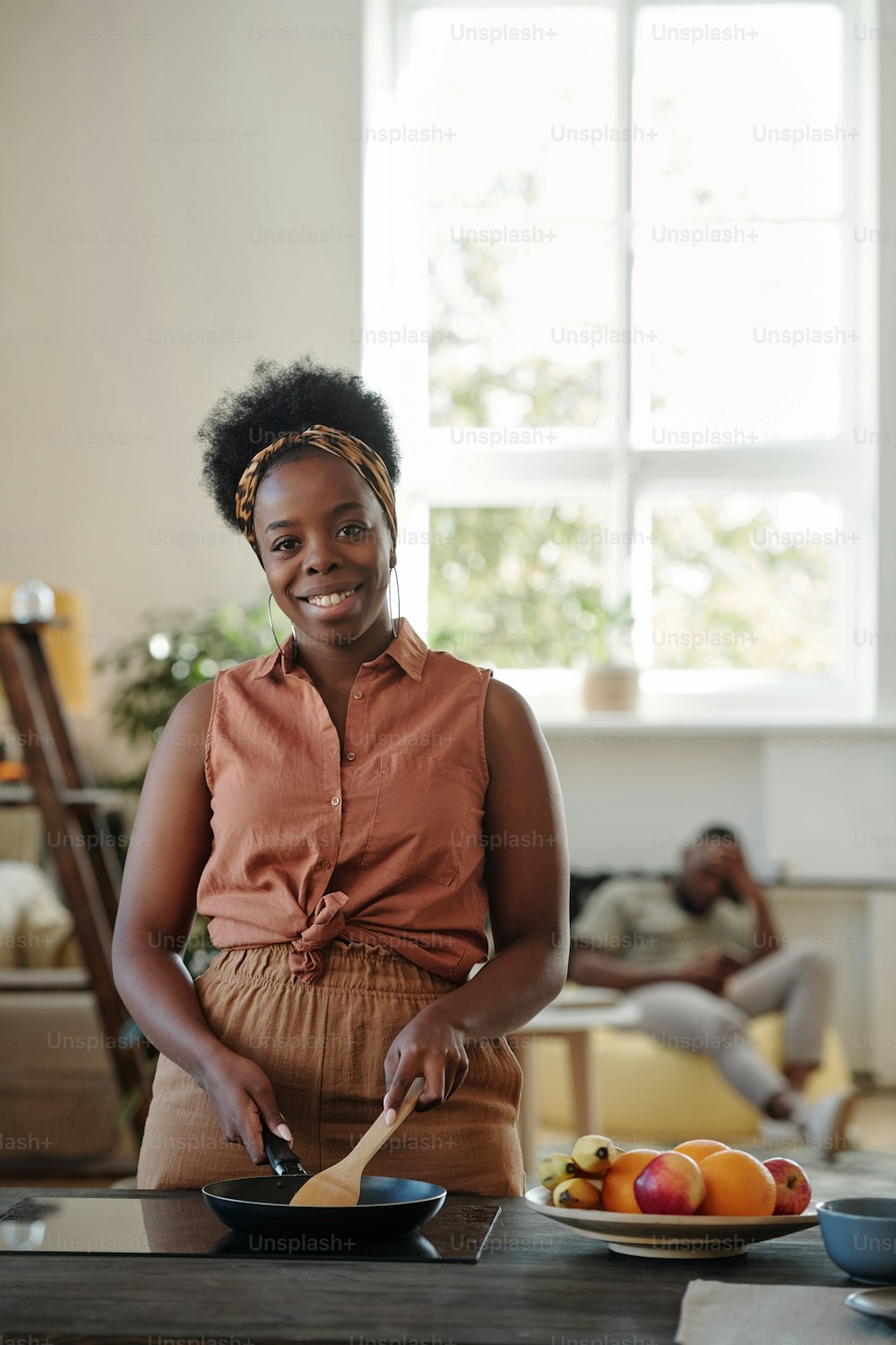 Jovem mulher sorridente de etnia africana olhando para você enquanto está de pé ao lado do fogão elétrico na cozinha e fritando algo para sua família