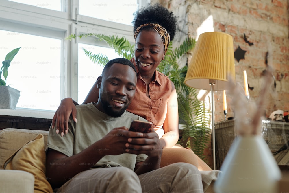 Giovane coppia africana gioiosa in abbigliamento casual che guarda lo schermo dello smartphone tenuto da un uomo sorridente mentre è seduto davanti alla telecamera contro la finestra