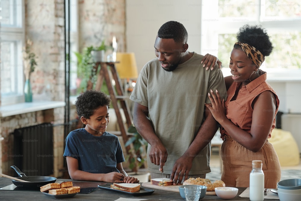 Young African man cutting bread for sandwiches by table in the kitchen for his affectionate wife and their adorable little son surrounding him