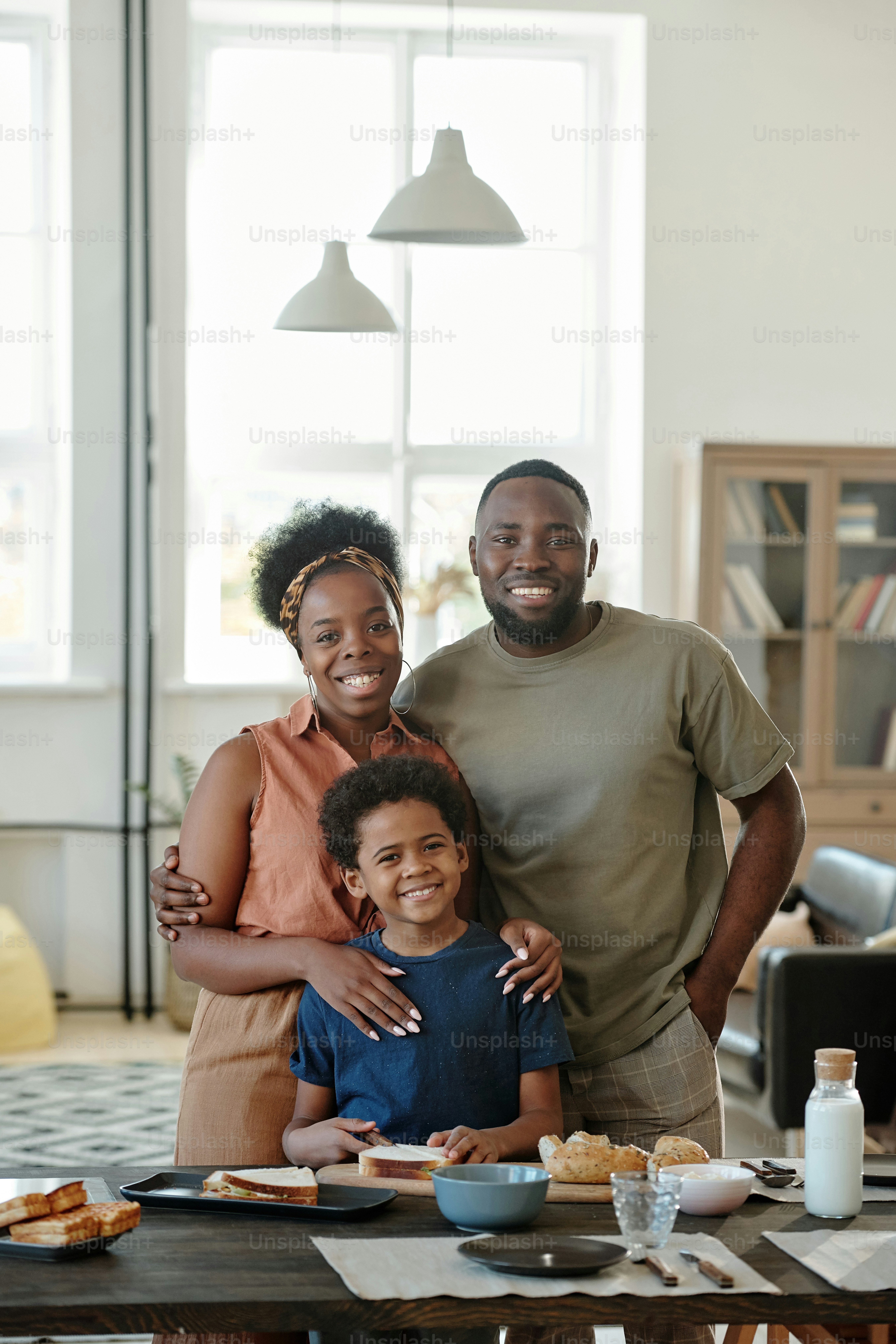 Joyful African family consisting of mother, father and cute little son standing by kitchen table in front of camera in home environment