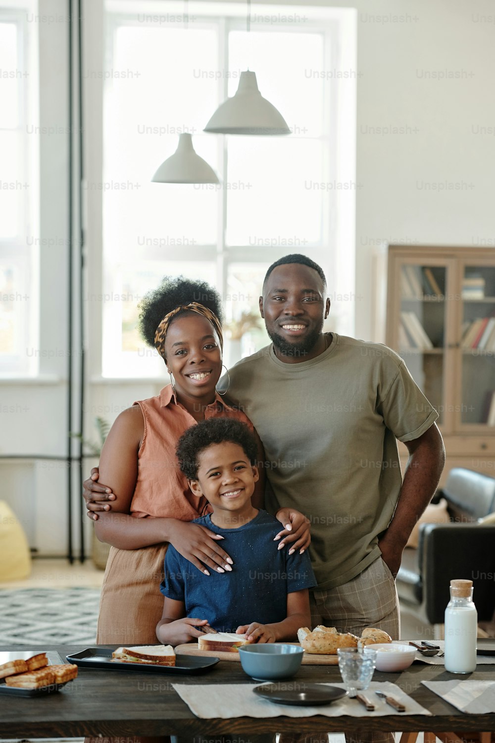 Joyful African family consisting of mother, father and cute little son standing by kitchen table in front of camera in home environment
