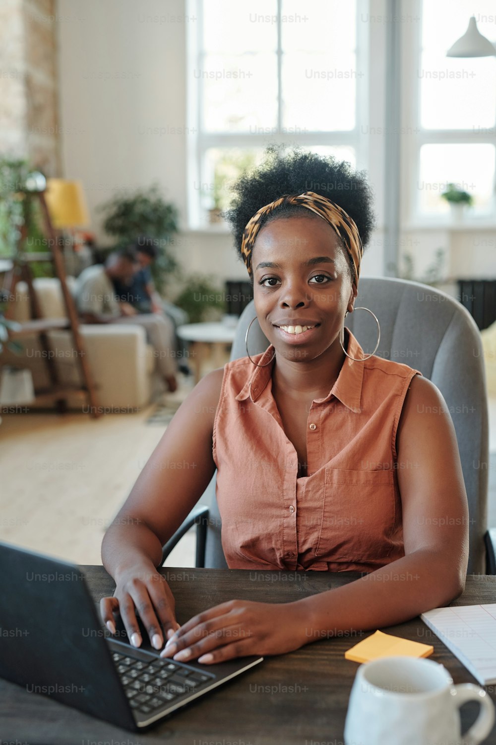 Giovane freelance africana di successo che ti guarda durante il lavoro davanti al laptop mentre è seduta in poltrona vicino al tavolo contro la sua famiglia