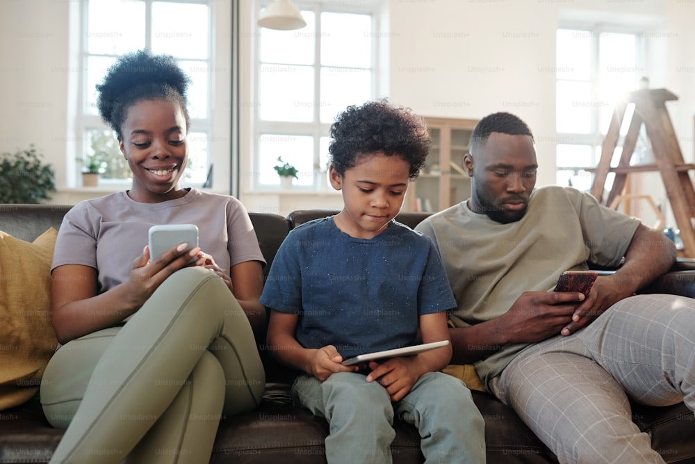 Young family of father, mother and cute little son using mobile gadgets while sitting in row on black leather couch in home environment