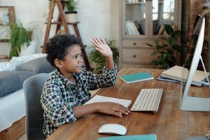 Heureux petit écolier assis près d’une table devant un écran d’ordinateur et faisant signe à son professeur avant le début de la leçon en ligne