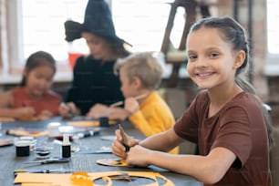 Portrait of smiling beautiful girl in brown tshirt sitting at table with craft supplies and making decoration at Halloween art class
