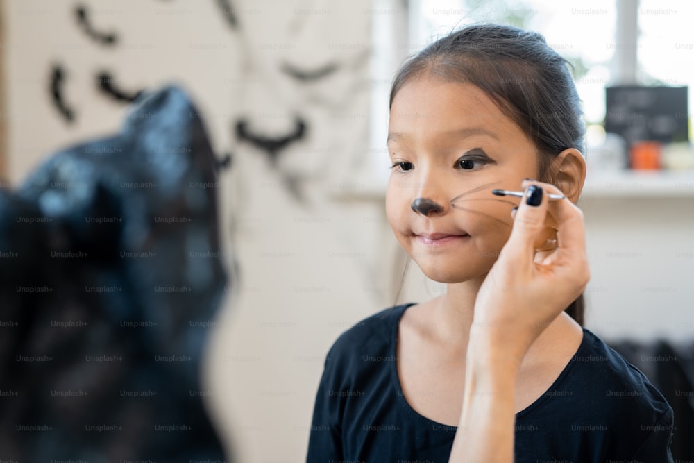 Vue par-dessus l’épaule de l’artiste peignant des moustaches sur le visage d’une fille asiatique tout en la préparant pour la fête d’Halloween