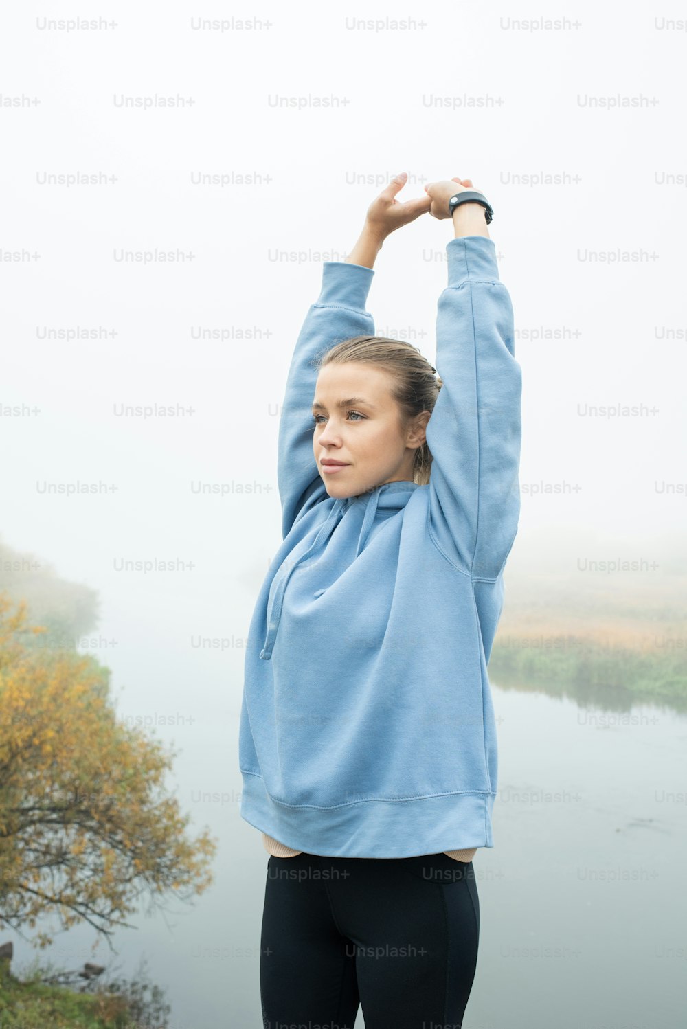 Giovane sportiva in forma in leggins neri e felpa blu con cappuccio che allunga le braccia sopra la testa mentre si esercita in un ambiente naturale in riva al fiume