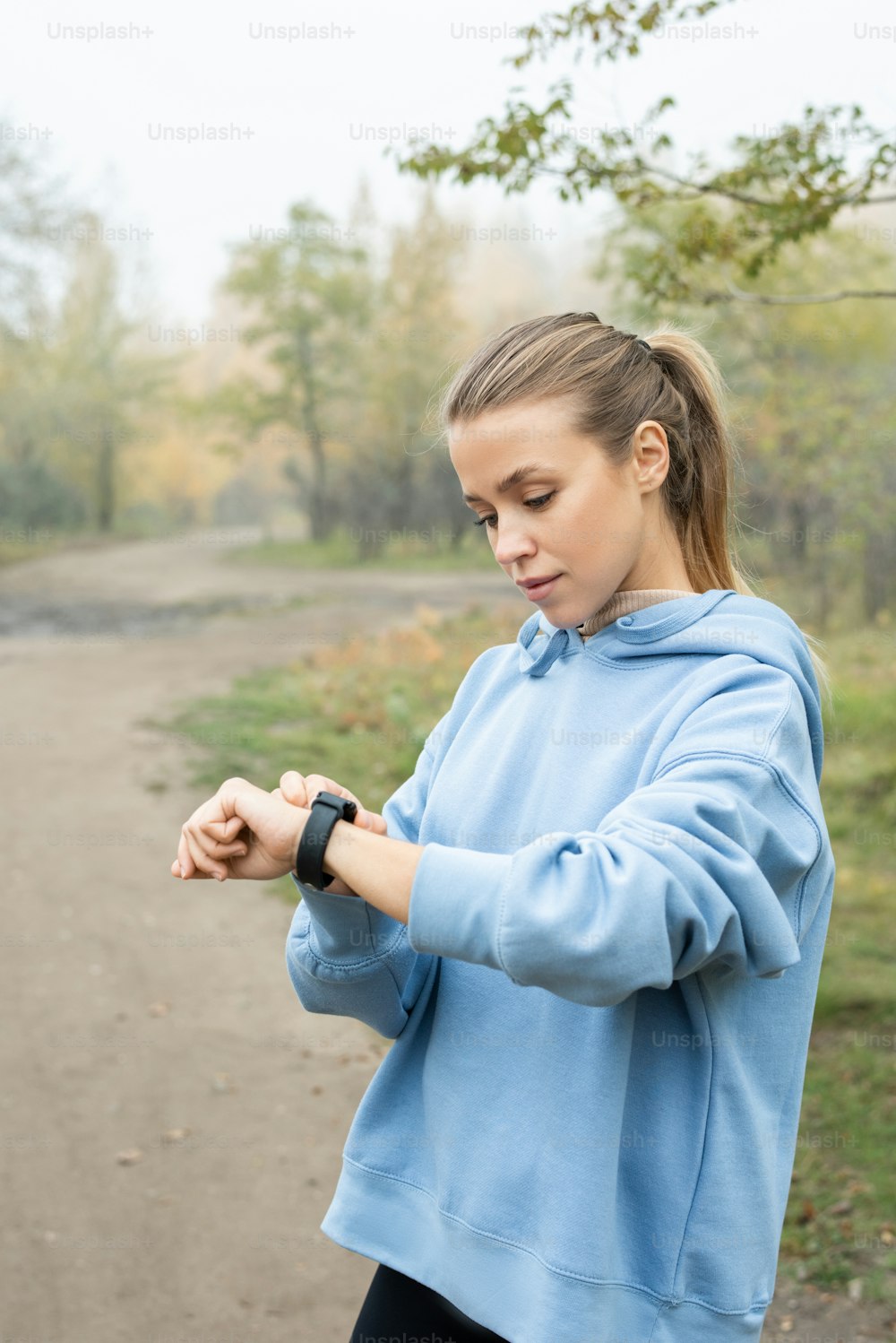 Giovane sportiva seria in felpa blu con cappuccio che guarda l'orologio da polso prima dell'allenamento mentre si trova davanti alla telecamera contro il sentiero della foresta