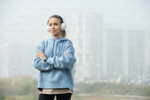 Young blond cross-armed sportswoman in white headphones and blue hoodie listening to music while standing in front of camera