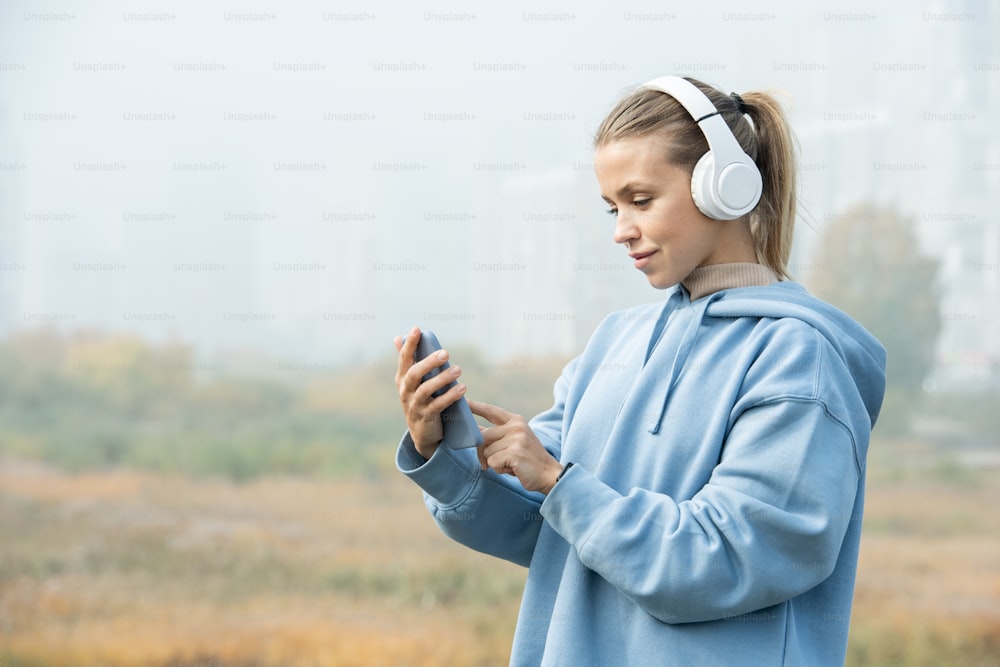Junge blonde Sportlerin in weißen Kopfhörern und blauem Kapuzenpulli hört Musik und zeigt auf den Bildschirm des Smartphones vor der Kamera