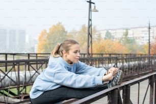 Young active sportswoman standing on bridge with stretched arms and right leg on metallic fence and exercising in the morning outdoors