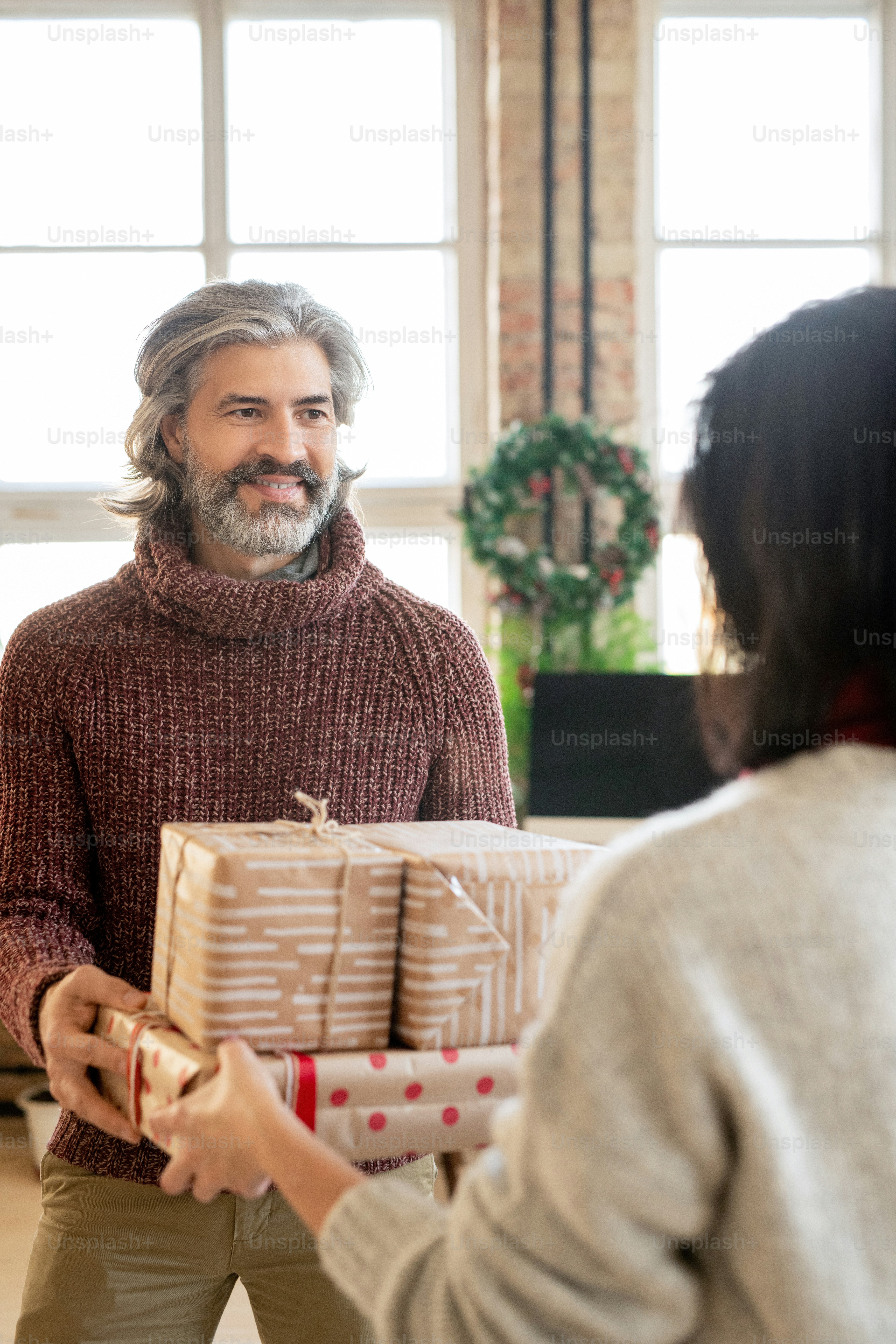 Uomo maturo felice che prende scatole regalo dalle mani di sua moglie mentre la guarda contro la ghirlanda di Natale sulla grande finestra nell'ambiente domestico