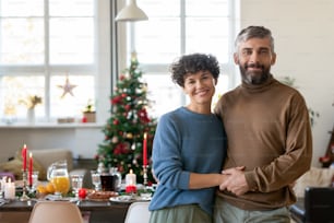 Young cheerful and affectionate couple in casualwear looking at you with smiles against served festive table prepared for family Christmas dinner
