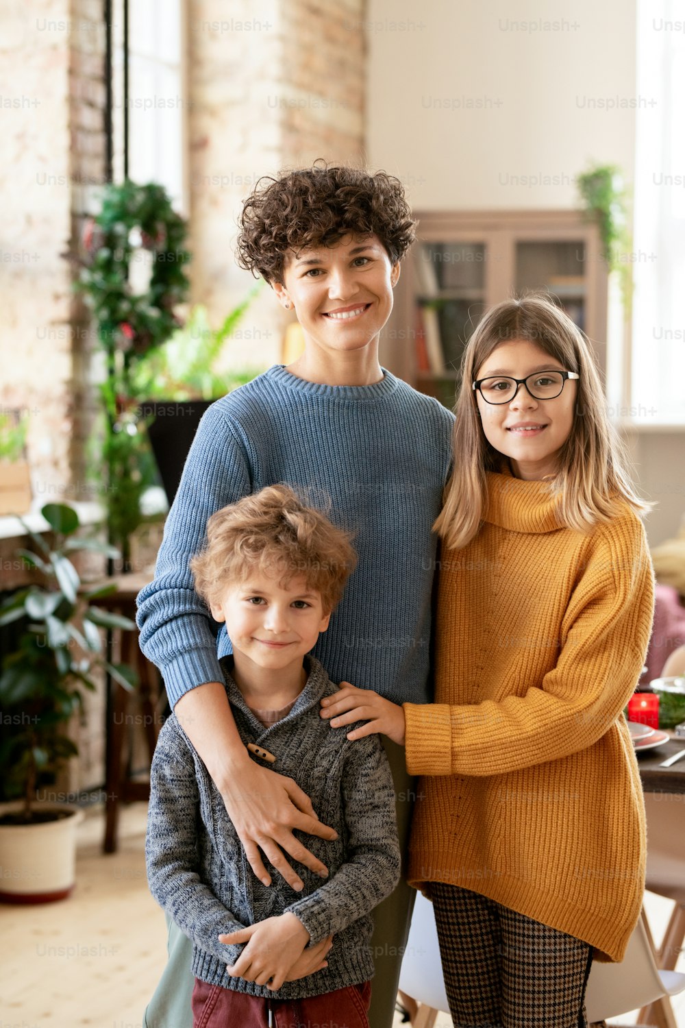Young cheerful brunette woman in casualwear embracing her two cute children while standing in front of camera on Christmas evening