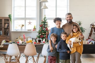 Joven familia grande y feliz de padres, tres niños lindos y su mascota de pie en la sala de estar el día de Navidad contra la mesa festiva servida