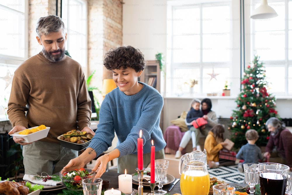 Happy husband and wife putting homemade salad, baked potatoes, drinks and other food on served festive table before family Christmas dinner