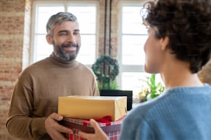 幸せなひげを生やした男性が妻に2つの贈り物を渡し、クリスマスリースとリビングルームの大きな窓を背景に笑顔で彼女を見ている
