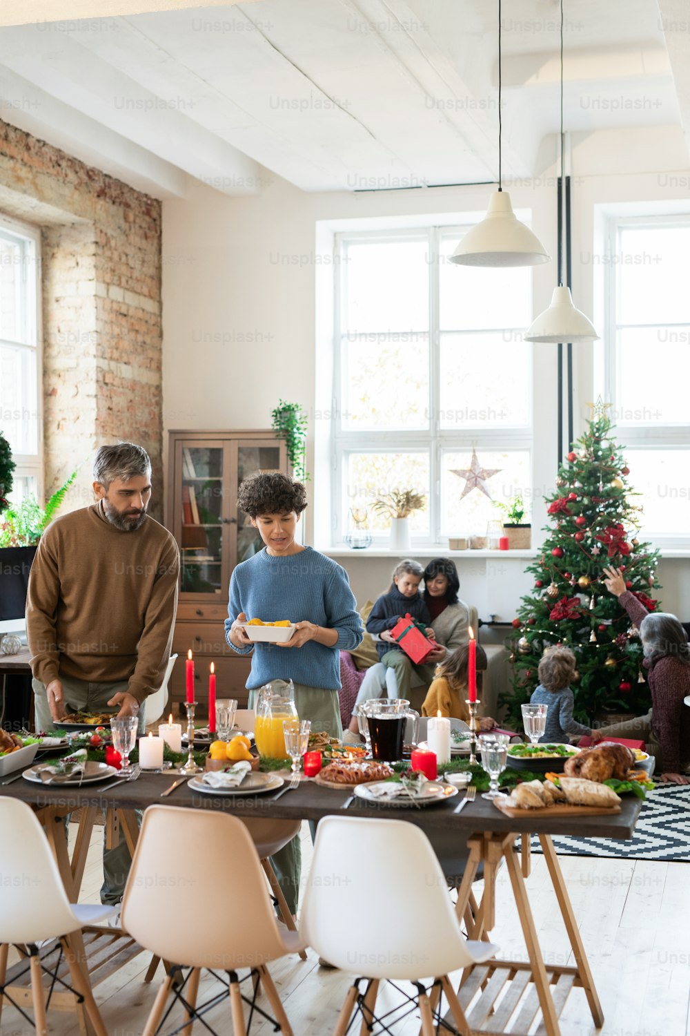 家族のクリスマスディナーの前に手作りの食べ物、飲み物、デザートを置きながら、お祝いのテーブルを待っている若い夫と妻