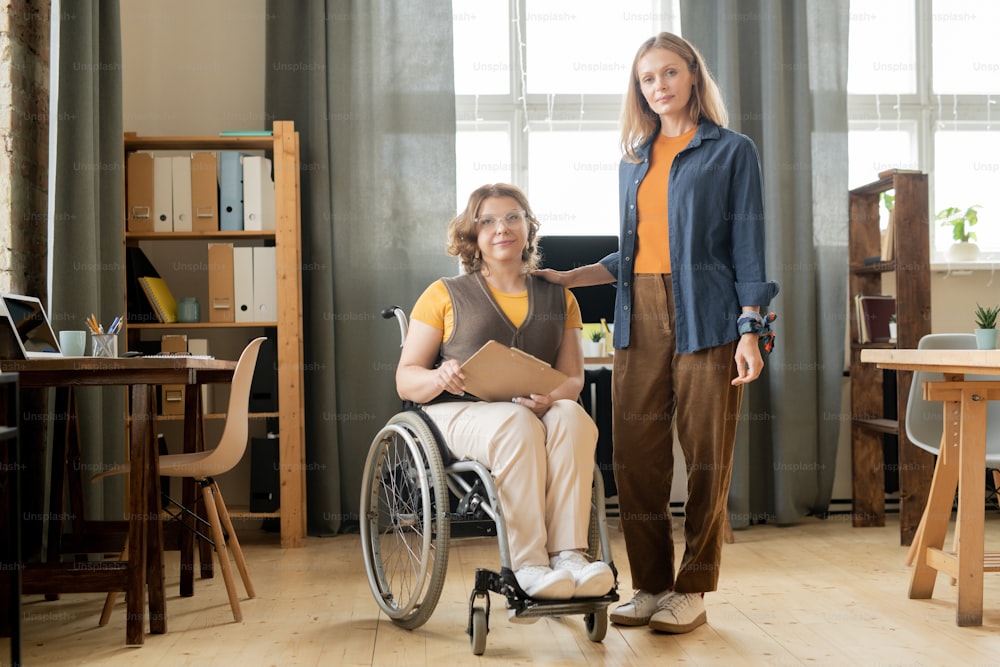 Young female manager and her disable colleague in casualwear standing in the center of office between workplaces against window and shelves