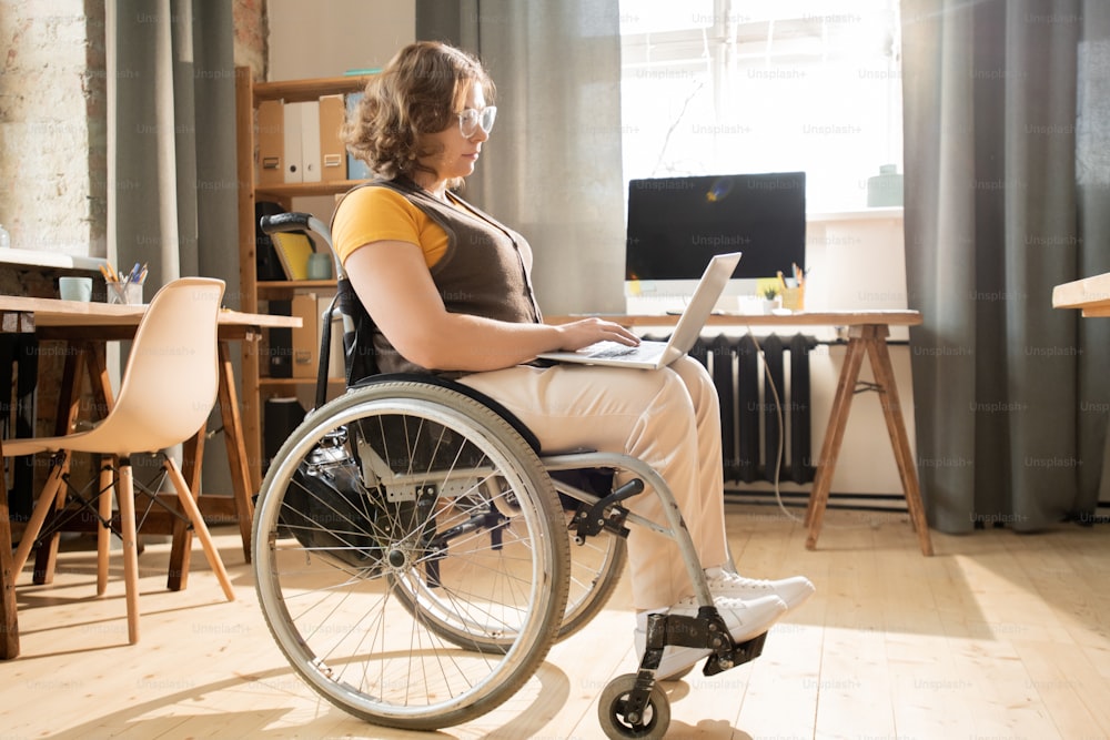 Vue latérale de la femme d’affaires handicapée en tenue décontractée tapant sur le clavier d’un ordinateur portable et regardant l’affichage tout en surfant sur le net dans un environnement domestique