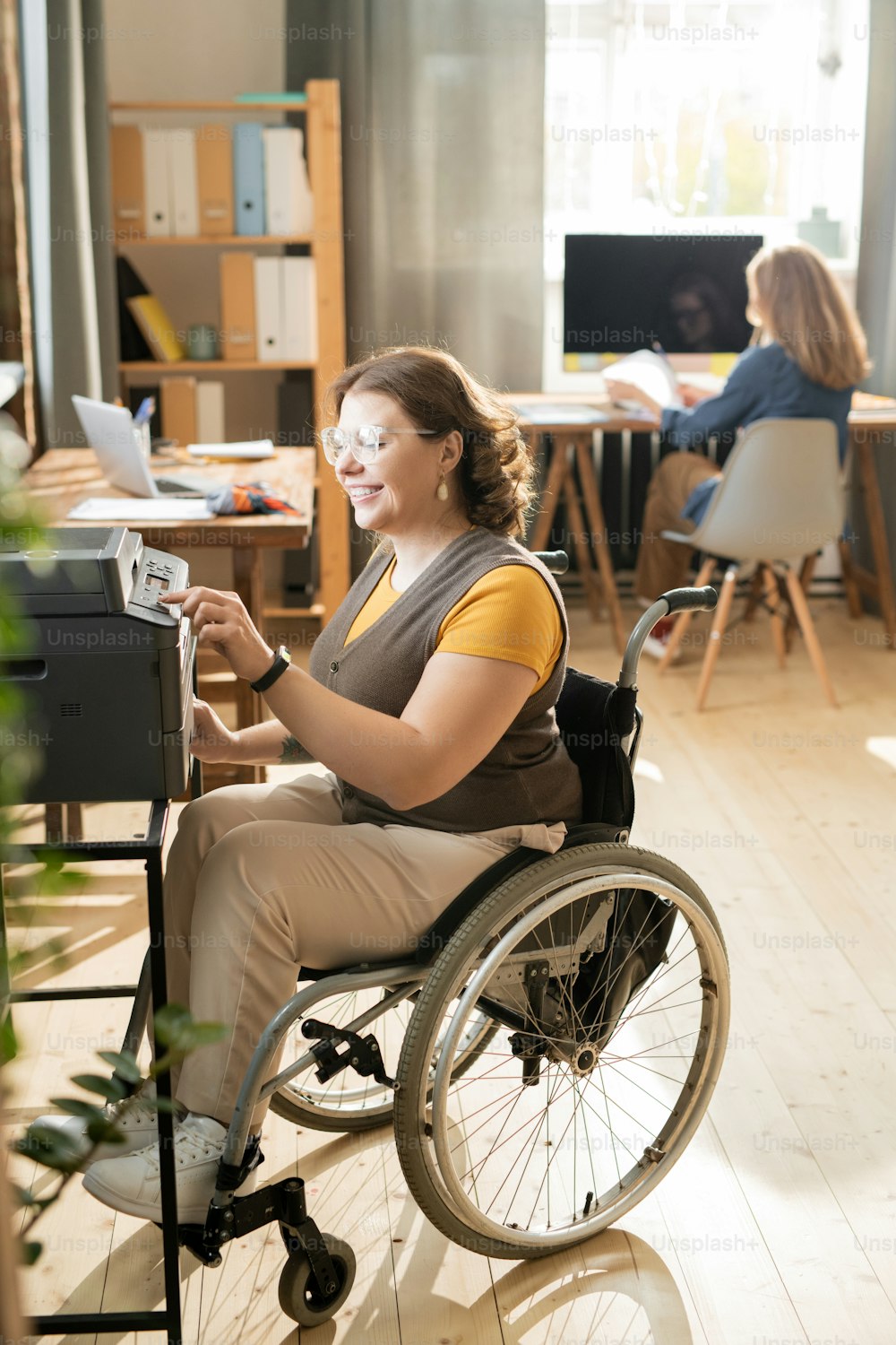 Jeune femme handicapée heureuse en fauteuil roulant assise près d’une machine Xerox et appuyant sur le bouton de démarrage tout en faisant des copies de documents au bureau