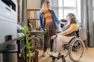 Junge blonde hübsche Frau in Freizeitkleidung, die ihre behinderte Freundin im Rollstuhl ansieht, während beide über Arbeitsmomente im häuslichen Zimmer diskutieren