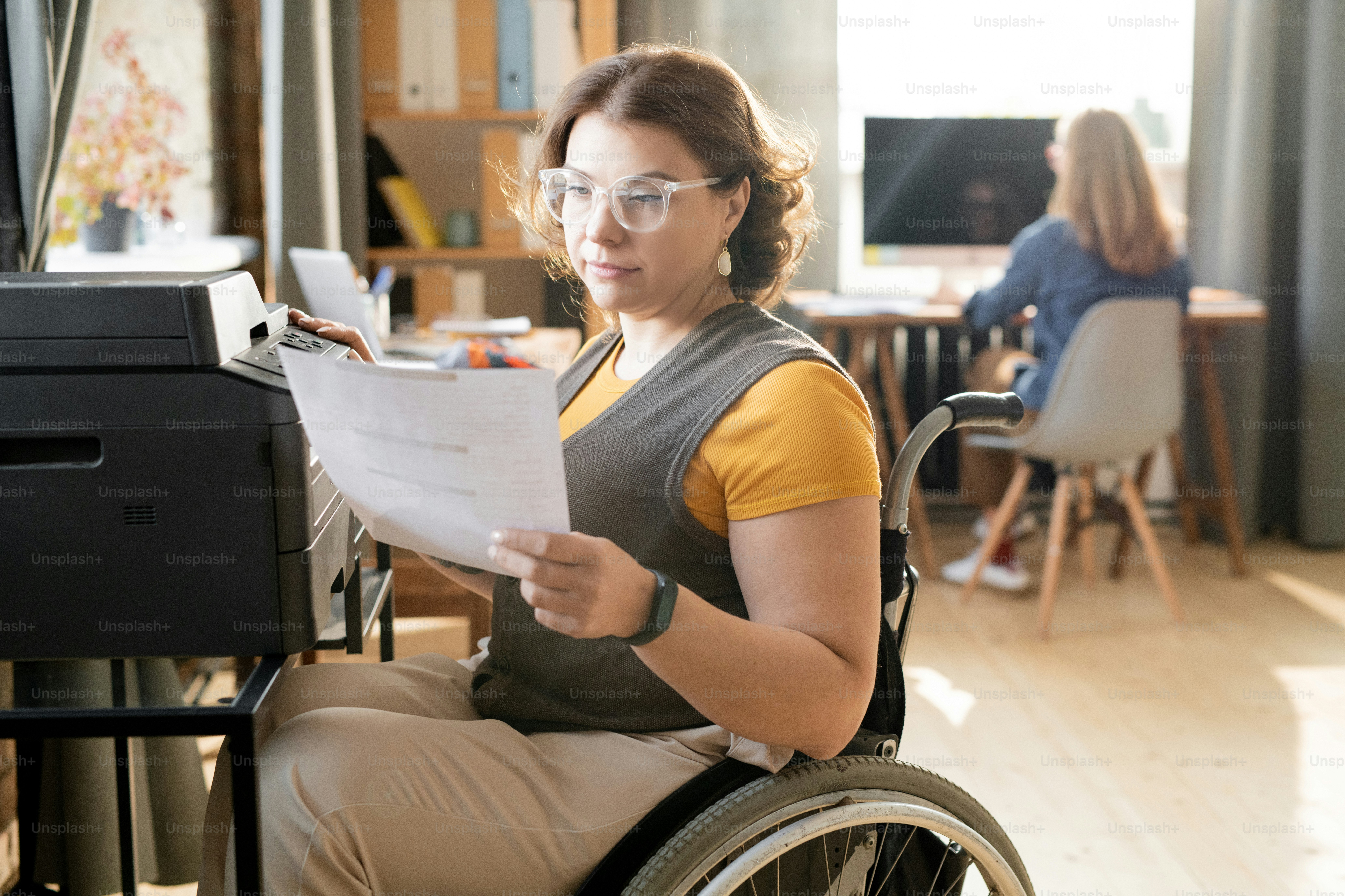 Young disable female office worker or secretary looking through paper while sitting by xerox machine and making copies against colleague