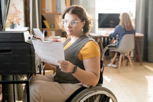 Young disable female office worker or secretary looking through paper while sitting by xerox machine and making copies against colleague