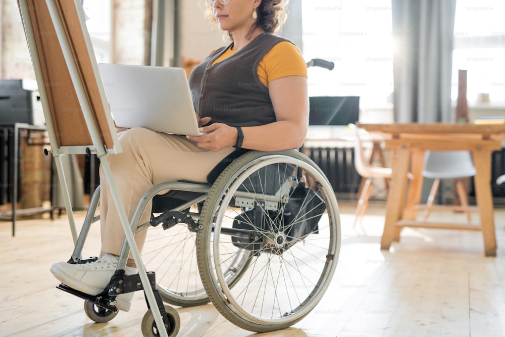 オフィスのホワイトボードの前でデザイナーやマネージャー向けのプレゼンテーションを準備しながら、ノートパソコンを使う車椅子の若い障害のある実業家
