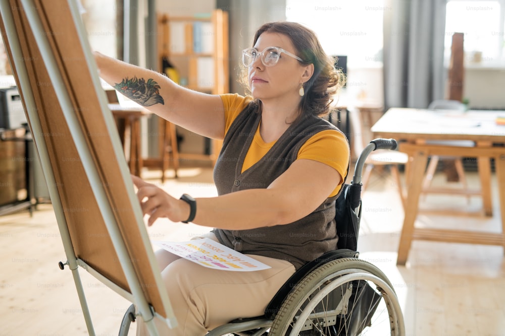 Young female business coach in casualwear sitting in wheelchair in front of whiteboard and preparing for seminar or presentation for designers