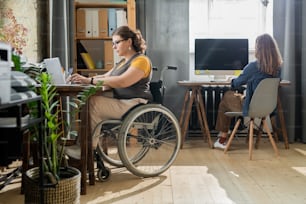 캐주얼웨어를 입은 젊은 장애인 사업가가 노트북 앞에 휠체어에 앉아 네트워킹을 하고 있는 동안 동료는 창가에서 일하고 있다