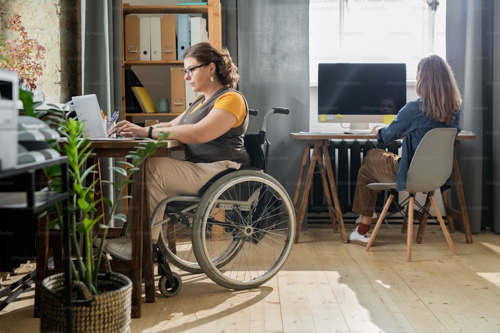 Young disable businesswoman in casualwear sitting in wheelchair in front of laptop and networking while her colleague working by window