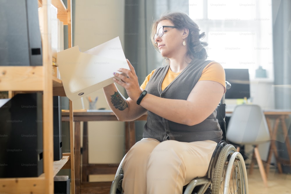 Joven trabajadora de oficina discapacitada mirando papeles en una de carpetas con documentos en el estante mientras busca un ejemplo de contrato